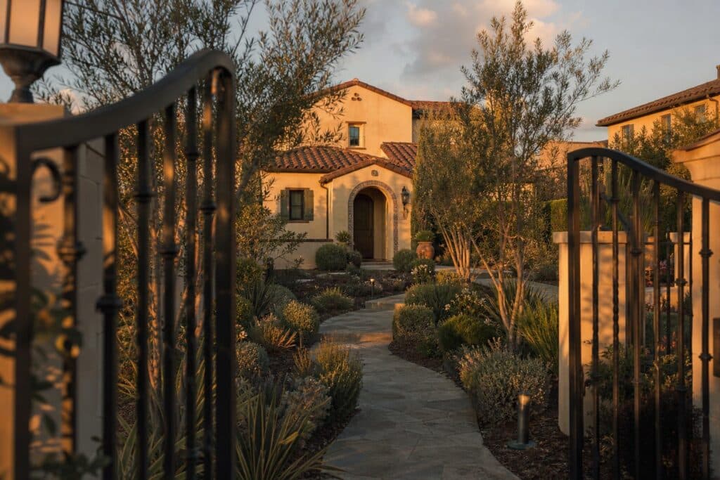 Gated Mediterranean-style home entrance with mature landscaping, stone pathway, and detached single-family residence in Porter Ranch Tuscany/Cortile