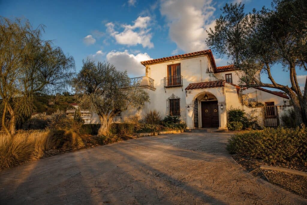 Vacant Spanish Colonial Revival home with terra cotta roof and mature trees in Granada Hills, afternoon sunlight illuminating aged stucco facade.
