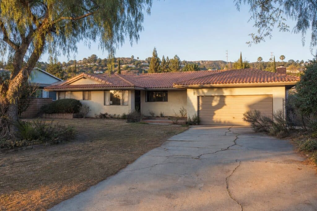 Weathered 1960s ranch home in Granada Hills with original stucco, tile roof, and mature pepper trees in afternoon light.